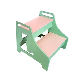 Könnte beinhalten: Ein zweistufiger Holzhocker mit hellgrüner Oberfläche und rosa gepolsterten Stufen. Der Hocker hat einen Ausschnitt in Form einer Katze auf der Vorderseite.