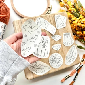 May include: A collection of Halloween-themed embroidery patterns, including bats, ghosts, and a dog holding a pumpkin. The designs are in gray on white fabric. A wooden embroidery hoop and orange yarn are in the background.