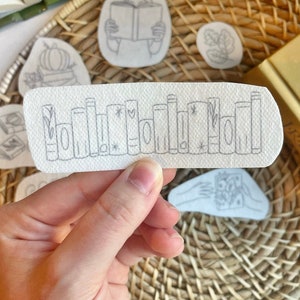 May include: A white sticker with a black line drawing of a row of books. The books are stacked vertically and have stars and hearts between them.
