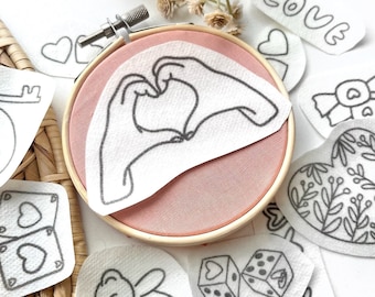 May include: A wooden embroidery hoop containing pink fabric with a white patch featuring a black line drawing of hands forming a heart shape. Surrounding the hoop are various white fabric patches with black line drawings of hearts, a teddy bear, dice, and the word LOVE.