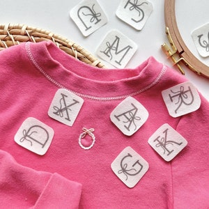 May include: A pink sweatshirt with white fabric letters sewn on it. The letters spell out the word "DREAM".