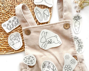 May include: A beige corduroy overall with wooden buttons, decorated with various white fabric patches featuring black line art illustrations of a rabbit, eggs, flowers, a chick, a carrot, bunny ears, and a bow, arranged on a woven surface.