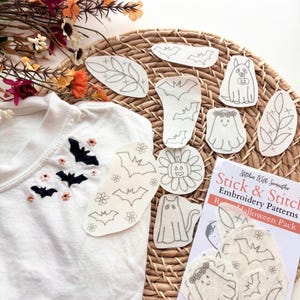 May include: A white t-shirt featuring black embroidered bats and small pink flowers, accompanied by a Stick & Stitch Embroidery Patterns Halloween Pack containing various adhesive fabric sheets with line art designs of ghosts, bats, and pumpkins.