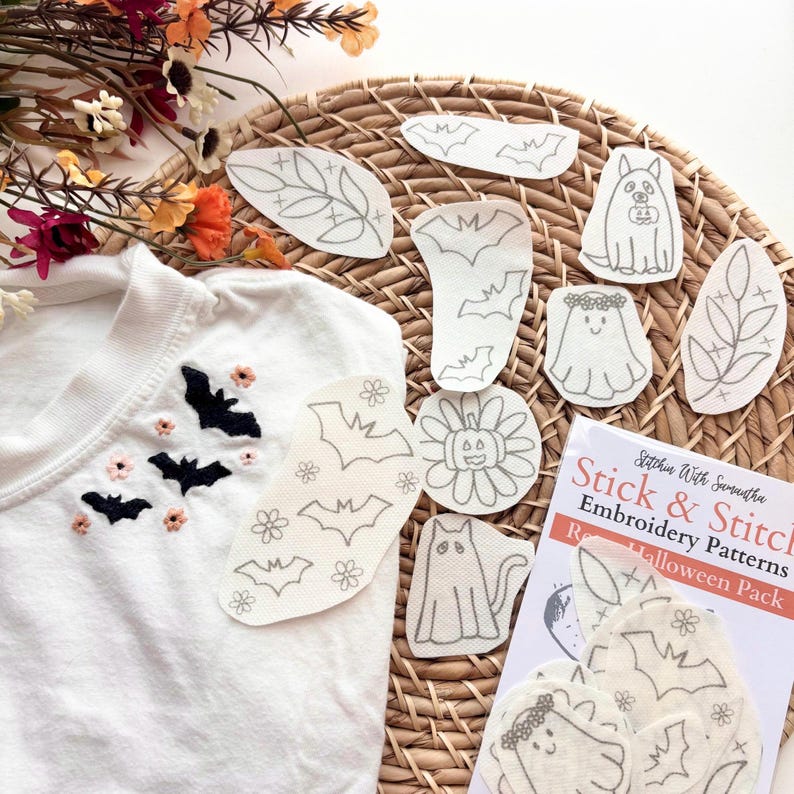 K&ouml;nnte beinhalten: Ein wei&szlig;es T-Shirt mit schwarzen gestickten Flederm&auml;usen und kleinen rosa Blumen, begleitet von Stick & Stitch Embroidery Patterns Halloween Pack B&ouml;gen mit Umrissen von Geistern, Flederm&auml;usen, Katzen und K&uuml;rbissen auf einer runden Flechtmatte.