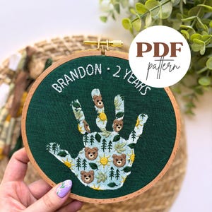 May include: Hand-embroidered hoop art with a green background. The design features a handprint filled with bears, trees, and suns. The text "BRANDON - 2 YEARS" is embroidered in white. A "PDF pattern" label is in the upper right.