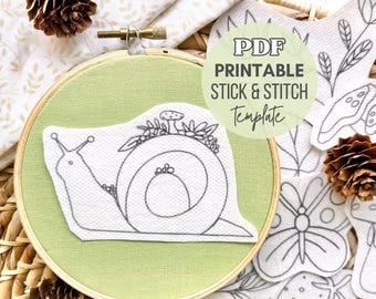 May include: A wooden embroidery hoop containing light green fabric with a white adhesive patch featuring a black line drawing of a snail with flowers on its shell. The text PDF PRINTABLE STICK & STITCH TEMPLATE is overlaid on the image.