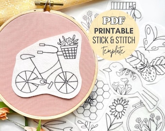 May include: A wooden embroidery hoop containing a dusty pink fabric with a white adhesive patch featuring a black line drawing of a bicycle with a flower basket. Surrounding the hoop are additional white stick and stitch embroidery patterns.