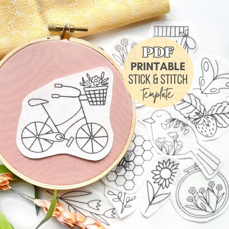 May include: A wooden embroidery hoop containing a dusty pink fabric with a white adhesive patch featuring a black line drawing of a bicycle with a flower basket. Surrounding the hoop are additional white stick and stitch embroidery patterns.