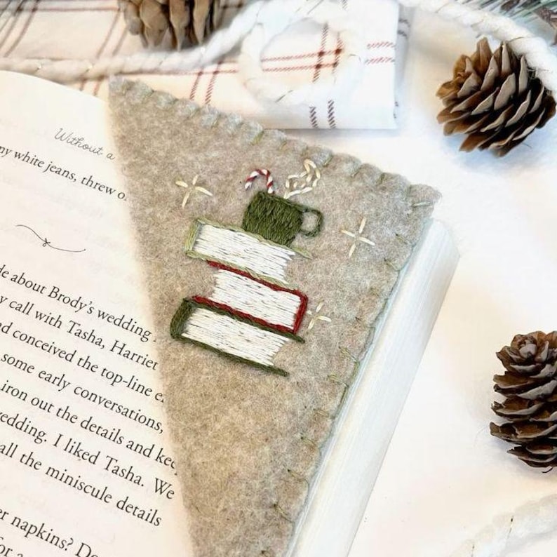 May include: A beige felt bookmark with embroidered details. The design features a stack of books with red spines, a green mug, and a candy cane. White cross-stitch accents the edges. The bookmark is placed in an open book.