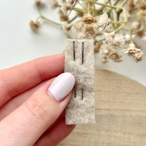 May include: A close-up of three silver needles on a beige felt pad. The needles are held in a hand with white nail polish.