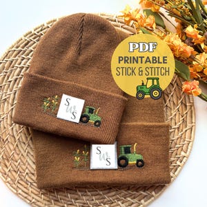 Peut inclure: Deux bonnets en tricot marron ornés de broderies représentant des tracteurs verts et des pieds de maïs. Chaque bonnet porte une petite étiquette carrée blanche avec les lettres S et W imprimées en noir. Un cercle jaune affiche le texte PDF PRINTABLE STICK & STITCH.