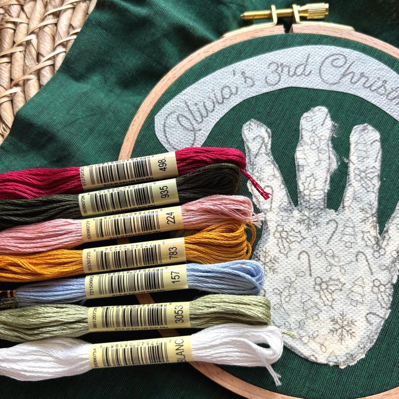 Puede incluir: Un bastidor de bordado con un fondo de tela verde. El bastidor presenta un dise&ntilde;o de huella de mano con elementos navide&ntilde;os y el texto "Olivia's 3rd Christmas". Varios carretes de hilo de bordar de colores est&aacute;n dispuestos debajo.