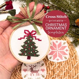 May include: A round wooden embroidery hoop containing white fabric with a cross-stitched green Christmas tree and red bow. A green ribbon and plaid fabric bow are tied at the top. Below are two additional cross-stitch patterns on paper.