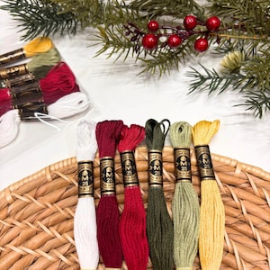 May include: Six skeins of DMC embroidery floss in white, dark red, bright red, dark green, light green, and yellow, each with a black and gold label marked 25 and 8m. Additional skeins are visible in the background with pine branches.