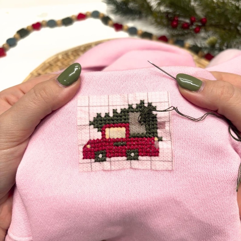 May include: A light pink fabric featuring a partially completed cross-stitch design of a red truck carrying a dark green Christmas tree. The embroidery is worked on a grid-patterned stabilizer, with a silver needle and dark thread visible.