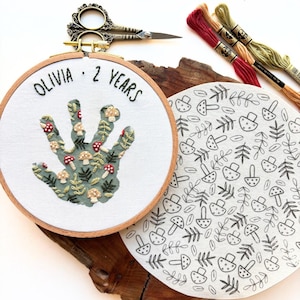 Peut inclure: Un tambour à broder en bois contenant un tissu blanc avec une forme d'empreinte de main remplie de champignons et de feuillages brodés, avec le texte OLIVIA 2 YEARS. À côté, un tissu avec un motif de champignons noirs et du fil à broder.