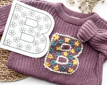 Könnte beinhalten: Ein langärmeliger Strickpullover in Mauve mit einem großen Buchstaben B auf der Vorderseite, der mit gelben und blaugrünen Blumenakzenten bestickt ist. Neben dem Pullover liegt eine weiße Karte mit einer Strichzeichnung des Buchstabens B.