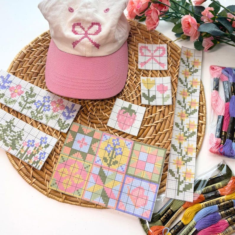 May include: A pink baseball cap with a bow design, surrounded by cross-stitch patterns of flowers, strawberries, and geometric shapes. Embroidery floss in various colors is also visible.