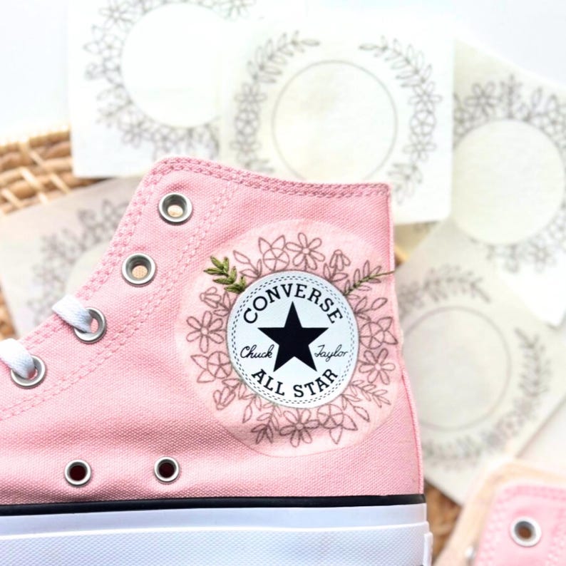 May include: A close-up of a pink Converse high-top sneaker with white laces and eyelets. The Converse logo is visible on the side, surrounded by a floral design. Several stickers with a floral wreath design are in the background.