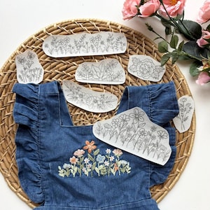 May include: A denim blue baby dress with ruffle sleeves and floral embroidery. Several floral embroidery patches are arranged around the dress on a woven basket. Pink flowers are in the background.