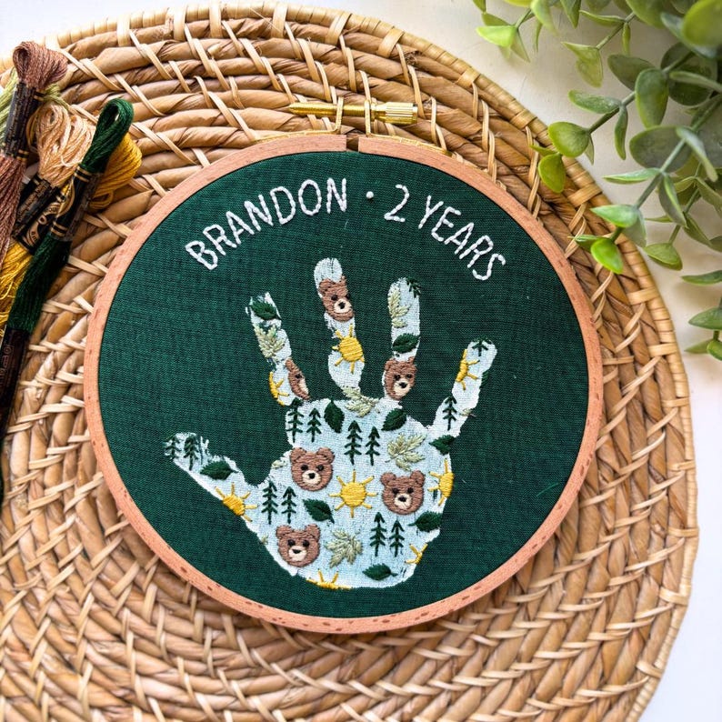 May include: Embroidered handprint art on a dark green background, featuring a handprint design with bears, trees, and suns. The text "BRANDON &middot; 2 YEARS" is stitched in white. The embroidery is set in a wooden hoop.