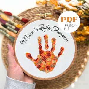 Könnte beinhalten: Gesticktes Stickrahmen-Kunstwerk mit einem Handabdruck-Design mit Herbstblättern und Kürbissen. Der Text "Mama's Little Pumpkin" ist über dem Handabdruck gestickt. Das Design befindet sich in einem Holzreifen.