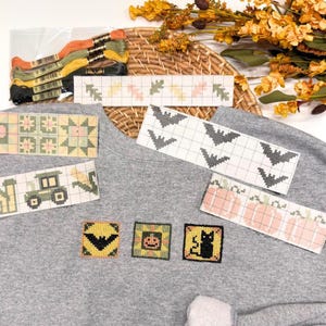 May include: A gray sweatshirt featuring three embroidered square patches of a bat, a pumpkin, and a black cat. Several cross-stitch pattern strips showing leaves, bats, pumpkins, and a tractor are arranged on the fabric with embroidery floss.