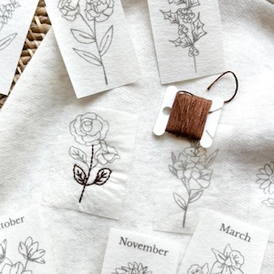 May include: Close-up of floral embroidery designs on small white fabric squares. A spool of brown thread sits nearby. Some squares have embroidered details, others are outlines. The word "October", "November", and "March" are visible.