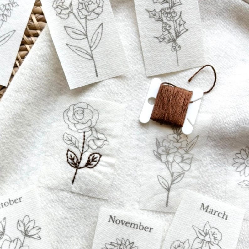 May include: Close-up of floral embroidery designs on small white fabric squares. A spool of brown thread sits nearby. Some squares have embroidered details, others are outlines. The word "October", "November", and "March" are visible.