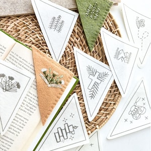 May include: A set of eight white felt triangle bookmarks with various embroidered designs, including a cross, a butterfly, a fence, a mountain range, a flower, and a tree. The bookmarks are arranged on a woven wicker surface.