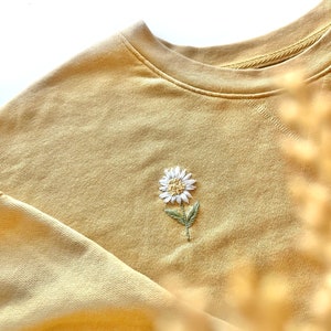 May include: A mustard yellow crewneck sweatshirt featuring a small, centered embroidery of a white daisy with a yellow center and green leaves. The garment has a ribbed neckline and long sleeves, showcasing a detailed floral stitch design.