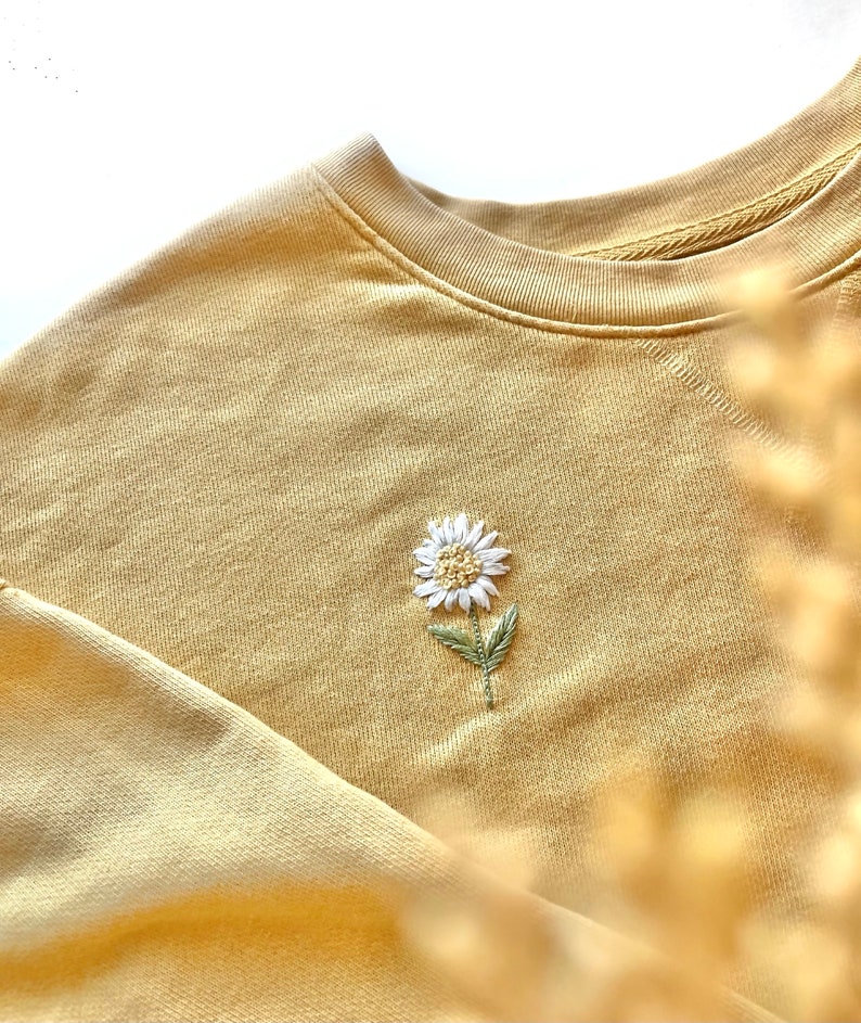 May include: A mustard yellow crewneck sweatshirt featuring a small, centered embroidery of a white daisy with a yellow center and green leaves. The garment has a ribbed neckline and long sleeves, showcasing a detailed floral stitch design.