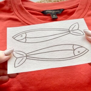 May include: A white sheet of paper with two black line drawings of fish. The fish are facing opposite directions. The paper is being held in front of a red shirt with a "Banana Republic" tag.