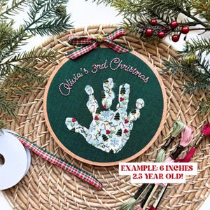 Puede incluir: Un bastidor de bordado con tem&aacute;tica navide&ntilde;a con un dise&ntilde;o de huella de mano. La huella est&aacute; llena de patrones festivos, incluyendo &aacute;rboles de Navidad y bastones de caramelo. El texto "Olivia's 3rd Christmas" est&aacute; bordado encima. El bastidor mide aproximadamente 15 cm de di&aacute;metro.