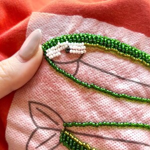 May include: A close-up of a red shirt with two green beaded fish appliques. The fish have white beads for eyes and a gold bead detail.