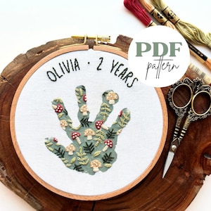 May include: An embroidered handprint design in a wooden hoop. The handprint is decorated with embroidered mushrooms and leaves in green, red, and beige. The text "OLIVIA - 2 YEARS" is stitched above. A PDF pattern is also available.