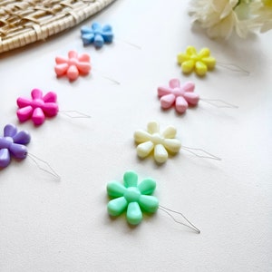 May include: A set of eight colorful flower-shaped threaders with metal needles. The threaders are in various pastel colors, including pink, purple, light blue, yellow, and green.