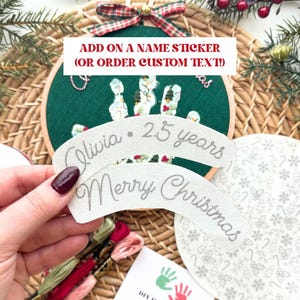 Puede incluir: Un aro bordado verde con huellas de manos y un lazo. El texto "ADD ON A NAME STICKER (OR ORDER CUSTOM TEXT!)" es visible. Una pancarta dice "Olivia &bull; 2.5 years" y otra "Merry Christmas".