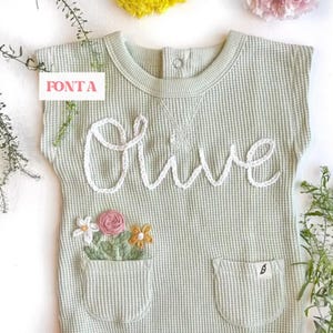 May include: A light green waffle knit onesie with a white embroidered name "Olive" and a pocket with embroidered flowers. The onesie has short sleeves and a round neckline.