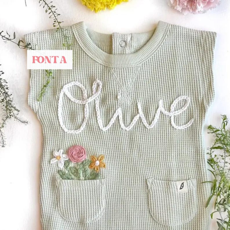 May include: A light green waffle knit onesie with a white embroidered name "Olive" and a pocket with embroidered flowers. The onesie has short sleeves and a round neckline.