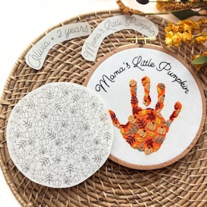 May include: An embroidered hoop with the text "Mama's Little Pumpkin" and a handprint design filled with autumn leaves and pumpkins. A separate round piece features a leaf and pumpkin pattern. Additional text reads "Olivia • 2 years".