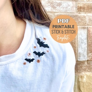 Puede incluir: Una camiseta blanca con un bordado de murciélago negro y flores rosas. La camiseta es una plantilla de pegar y coser, con el texto "PDF Printable Stick & Stitch Template" en un círculo.