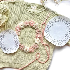 May include: A light green knitted sweater with a pink and white flower embroidery design. The sweater has a round neckline and long sleeves. There are two white circles with black outlines of flowers on the sweater.