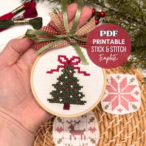 Peut inclure: Un tambour à broder circulaire en bois contenant un tissu blanc avec un sapin de Noël vert foncé brodé au point de croix, orné de décorations rouges et d'un nœud rouge. Un ruban vert est noué au sommet. Texte : PDF PRINTABLE STICK & STITCH Template.