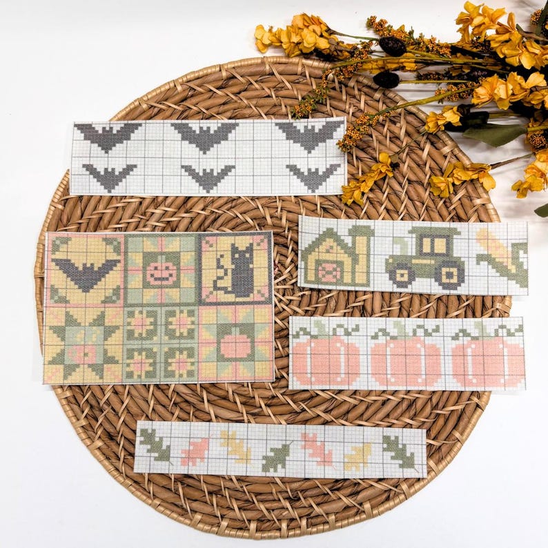 May include: A collection of cross-stitch pattern charts printed on paper, featuring seasonal motifs including bats, a black cat, a pumpkin, a tractor, a barn, and autumn leaves in shades of orange, green, yellow, and gray on a grid background.