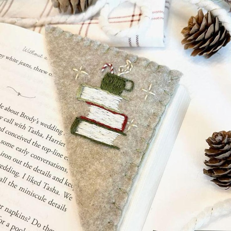 May include: A beige felt bookmark with an embroidered design of a stack of books, a mug of hot cocoa with a candy cane, and a few small white crosses. The bookmark is placed on an open book.