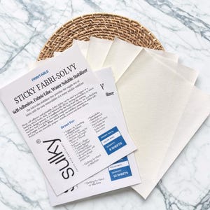 May include: White sheets of printable, self-adhesive, fabric-like, water-soluble stabilizer, with the text "STICKY FABRI-SOLVY" and "SULKY" printed on the sheets. The sheets are on a marble surface with a woven basket.