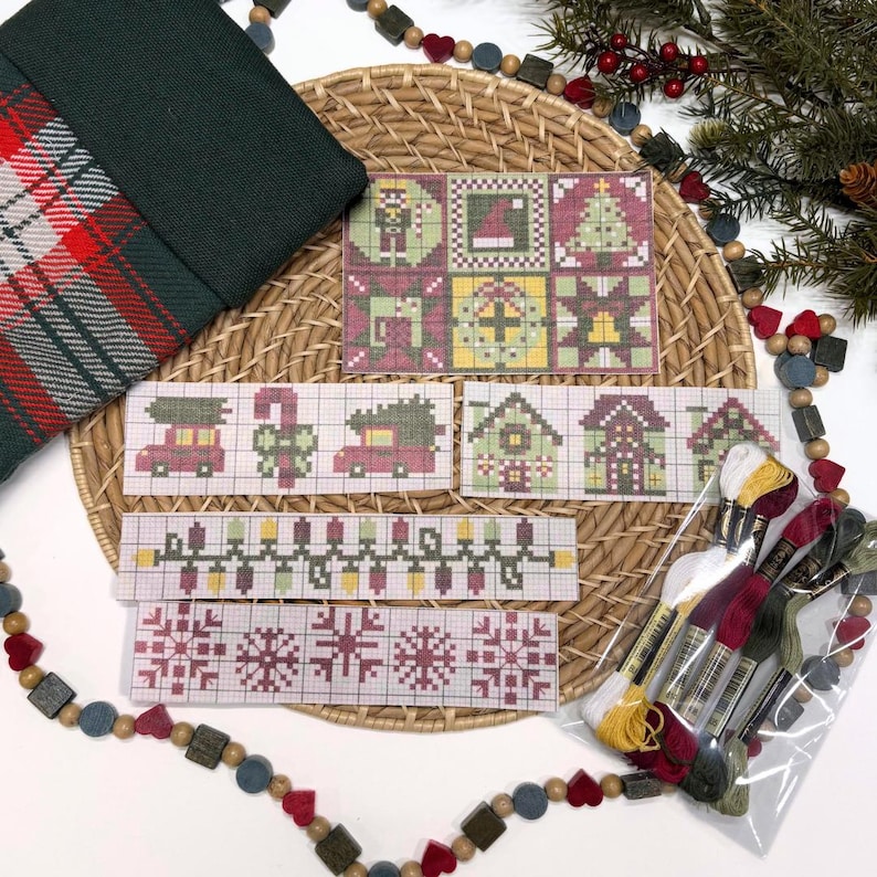May include: A cross-stitch embroidery kit featuring multiple holiday-themed patterns on grid paper, including snowflakes, Christmas trees, houses, and light strings, accompanied by a clear plastic bag containing assorted colored embroidery floss.