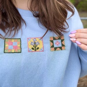 May include: Light blue sweatshirt with three embroidered square patches. Each patch features a different floral or geometric design in pastel colors. The sweatshirt is made of a soft material.