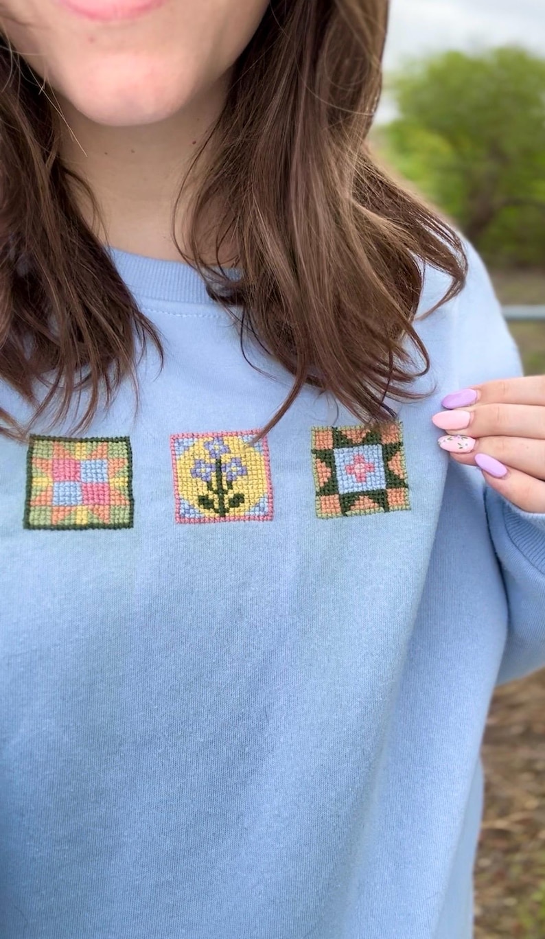 May include: Light blue sweatshirt with three embroidered square patches. Each patch features a different floral or geometric design in pastel colors. The sweatshirt is made of a soft material.